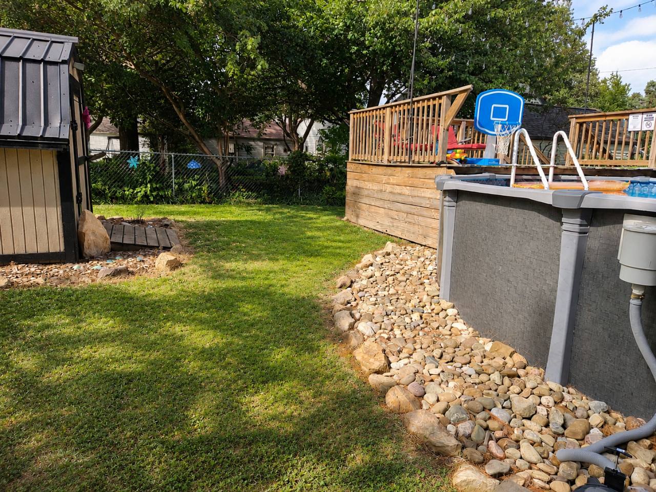 Above-ground pool beside its wooden deck, with a pool basketball hoop and landscaped stone border