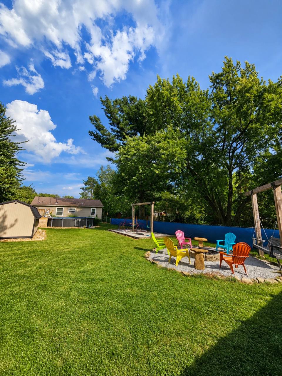Spacious backyard with colorful chairs around a fire pit, swings and an above-ground pool in the distance
