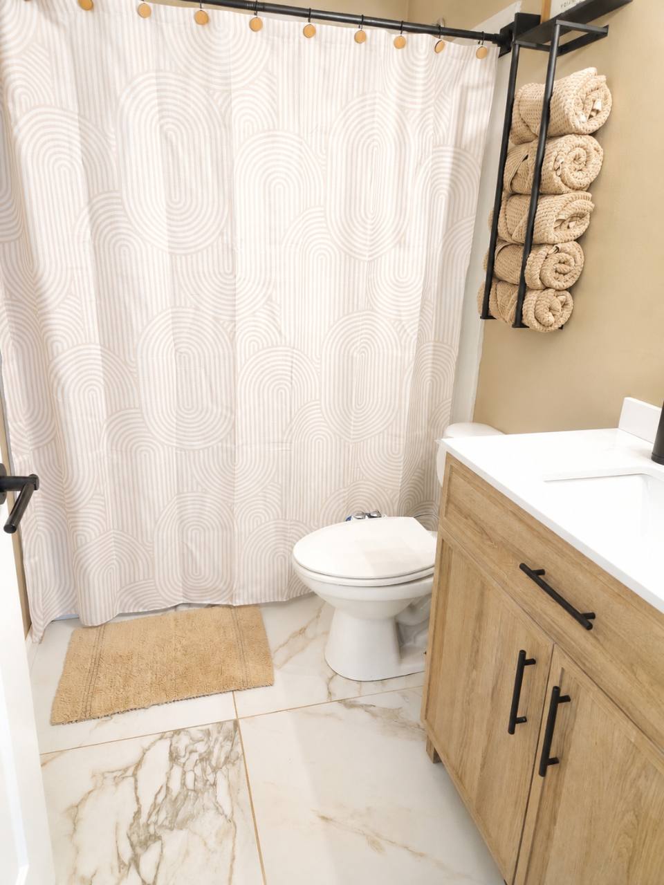 Bathroom with patterned shower curtain, wood vanity, toilet and rolled towels on a wall rack