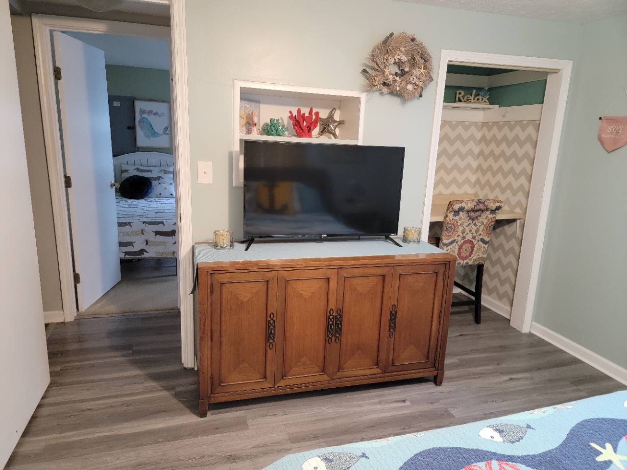 Bedroom television on a wooden cabinet beside a makeup nook and a doorway toward the bed