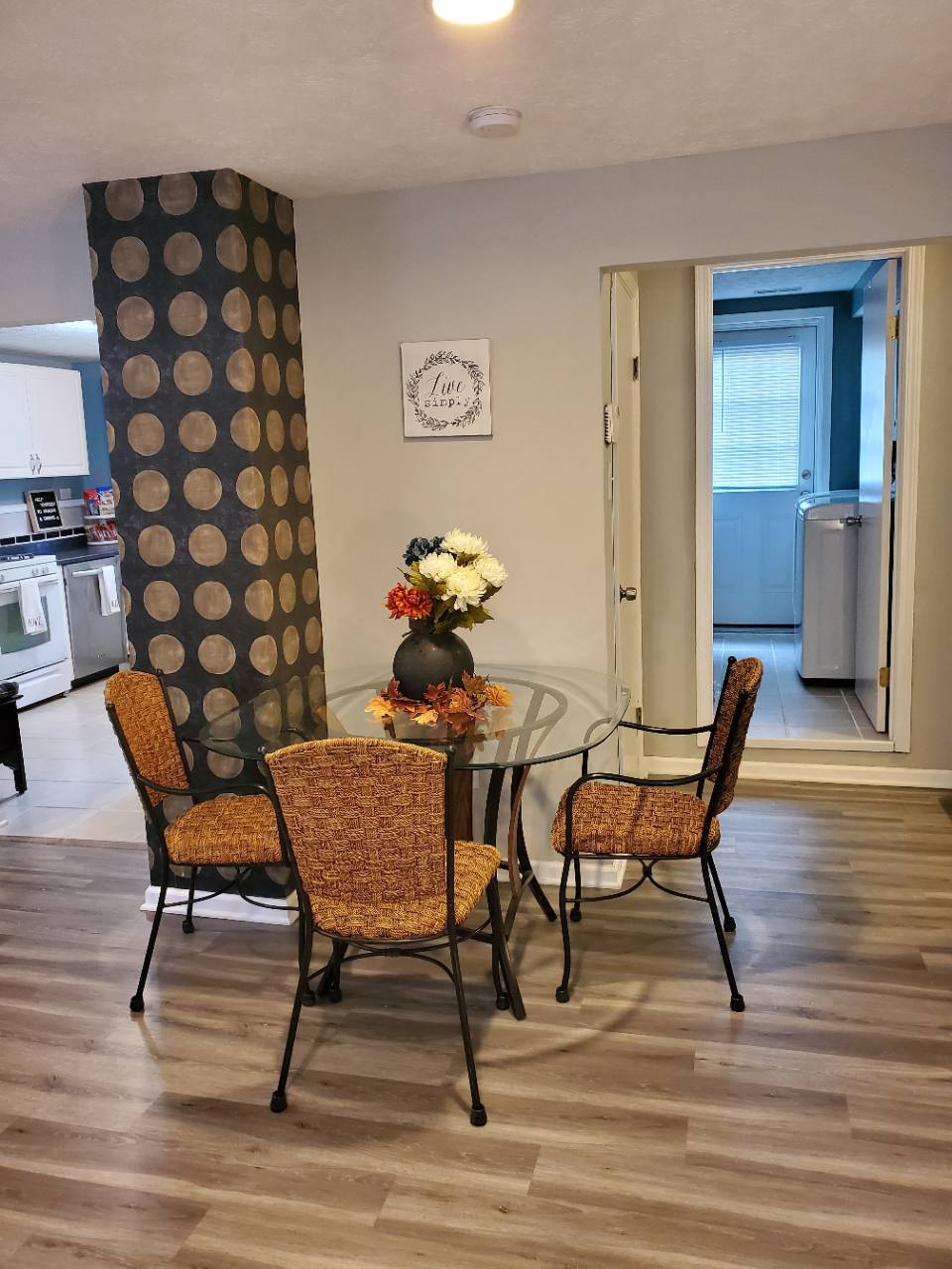 Round glass dining table with woven chairs beside doorways to the kitchen and bathroom laundry area