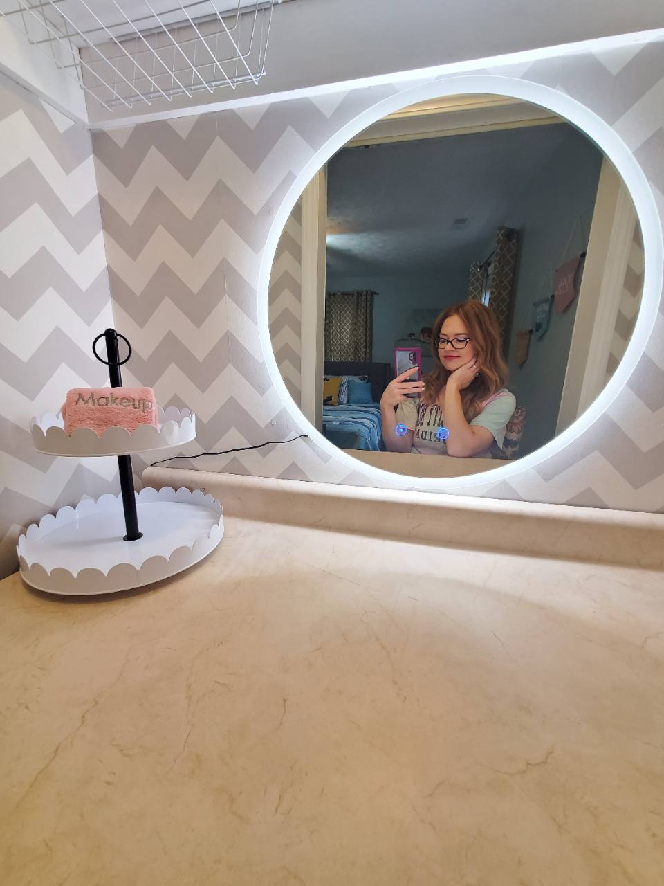 Makeup countertop with illuminated round mirror, chevron-patterned walls and the photographer reflected in the mirror